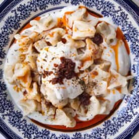 serving platter showcasing manti dumplings topped with creamy garlicky yogurt and bright red tomato butter sauce, garnished with dried mint and Aleppo pepper