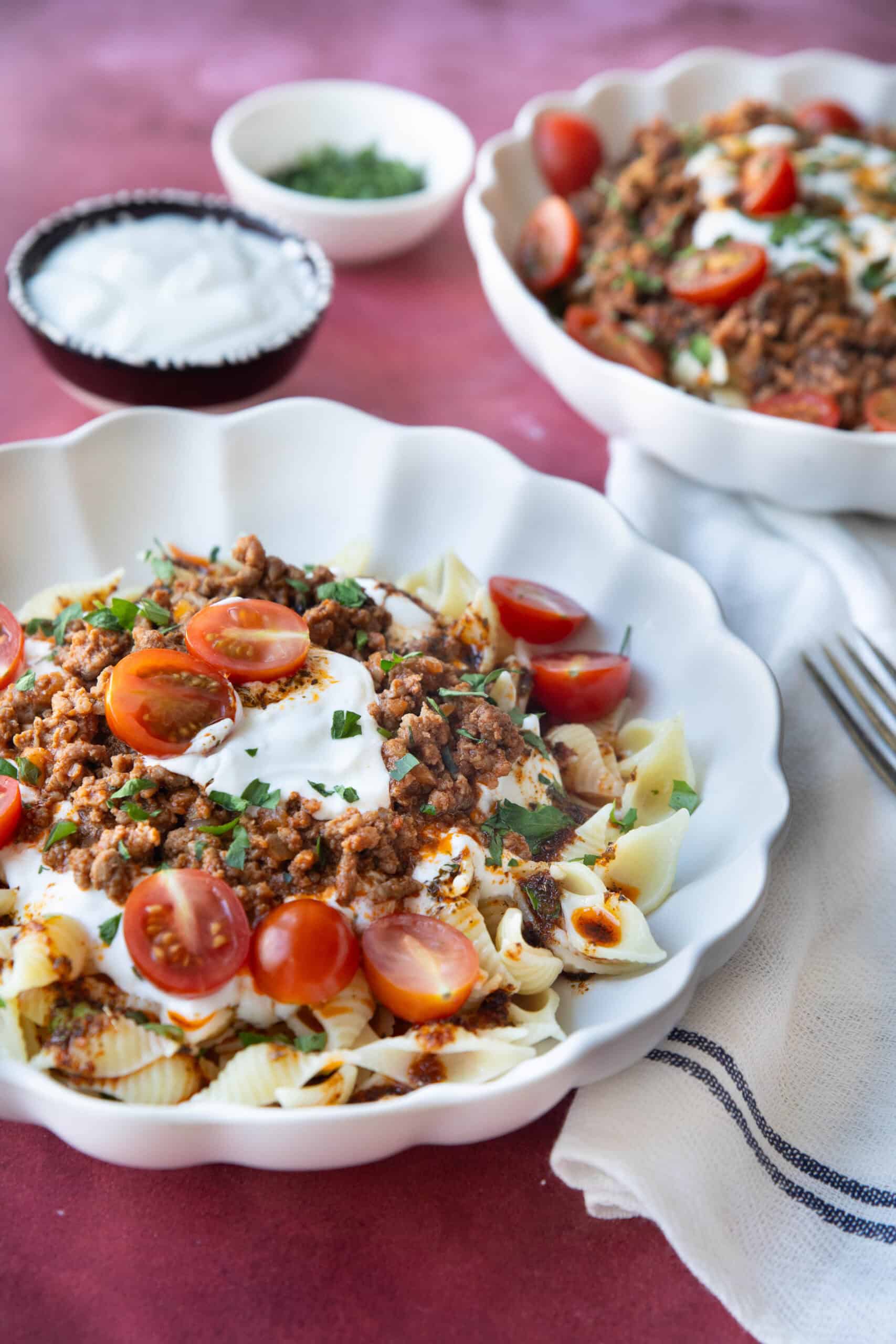Turkish Pasta with Spiced Butter Two plates of Turkish pasta served on a white ceramic plate, topped with spiced ground beef, a generous drizzle of creamy garlic yogurt, and a golden paprika butter sauce, garnished with fresh parsley and cherry tomatoes.