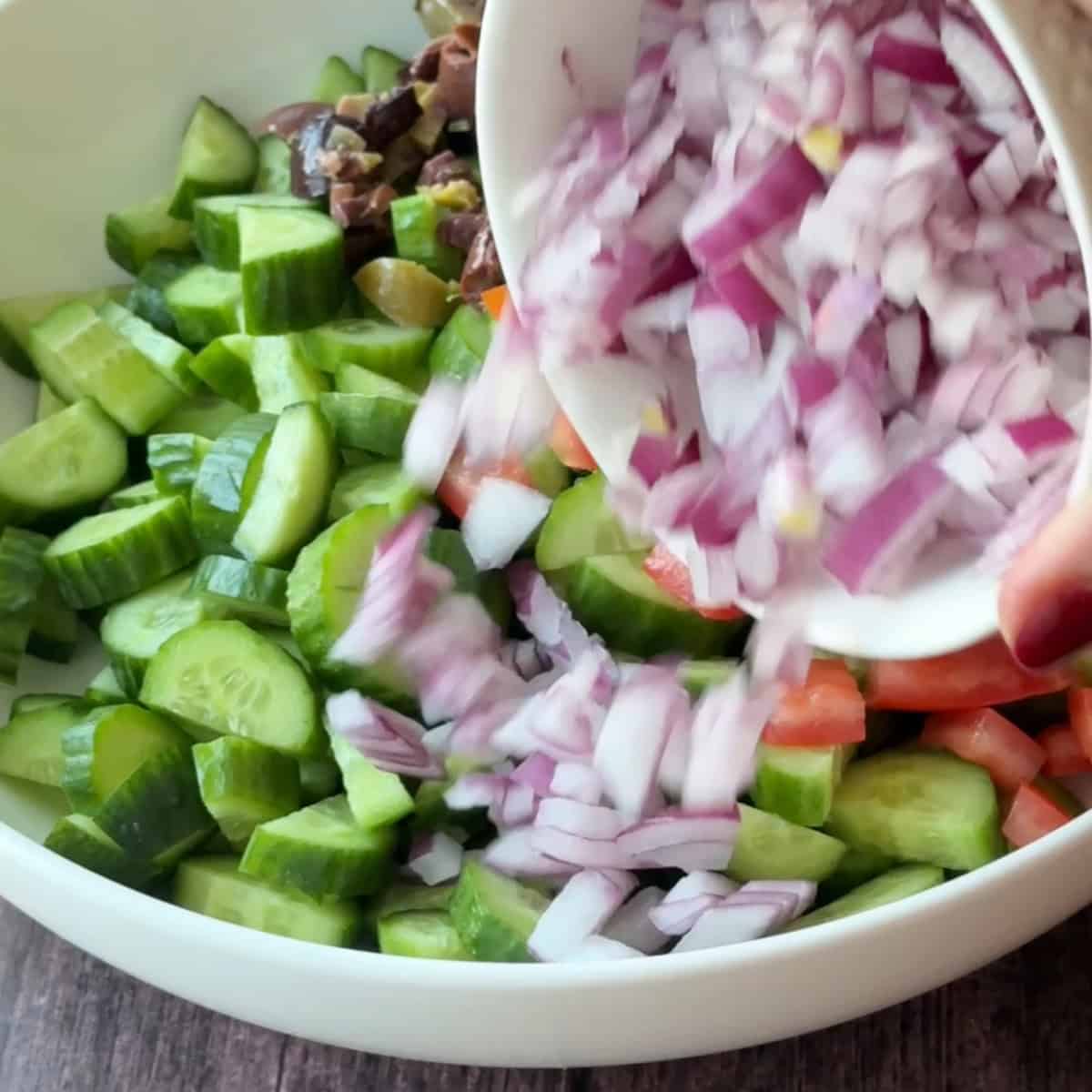 How to Make Mediterranean Cucumber Salad adding chopped red onions over cucumber, tomatoes, and olives