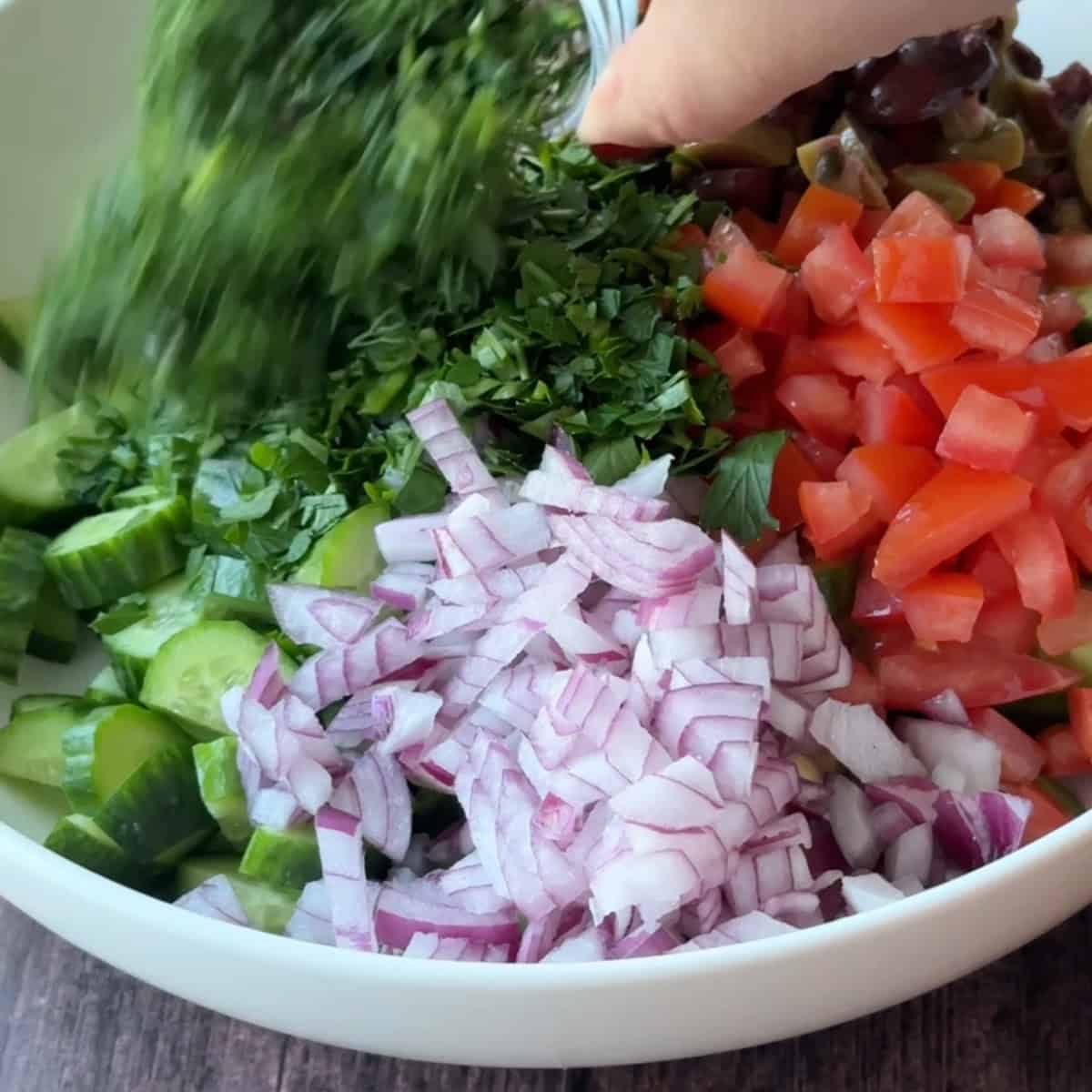 How to Make Mediterranean Cucumber Salad adding chopped fresh parsley over cucumber, tomatoes, red onions, and olives