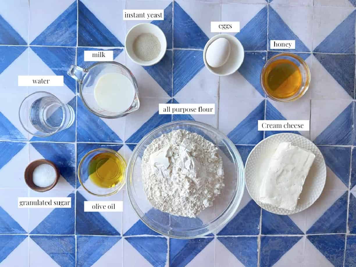 khaliat Nahal Ingredients honeycomb bread ingredients, including flour, yeast, warm milk, warm water, olive oil, sugar, salt, cream cheese, an egg, sesame seeds, nigella seeds, and honey, neatly arranged on a kitchen countertop.