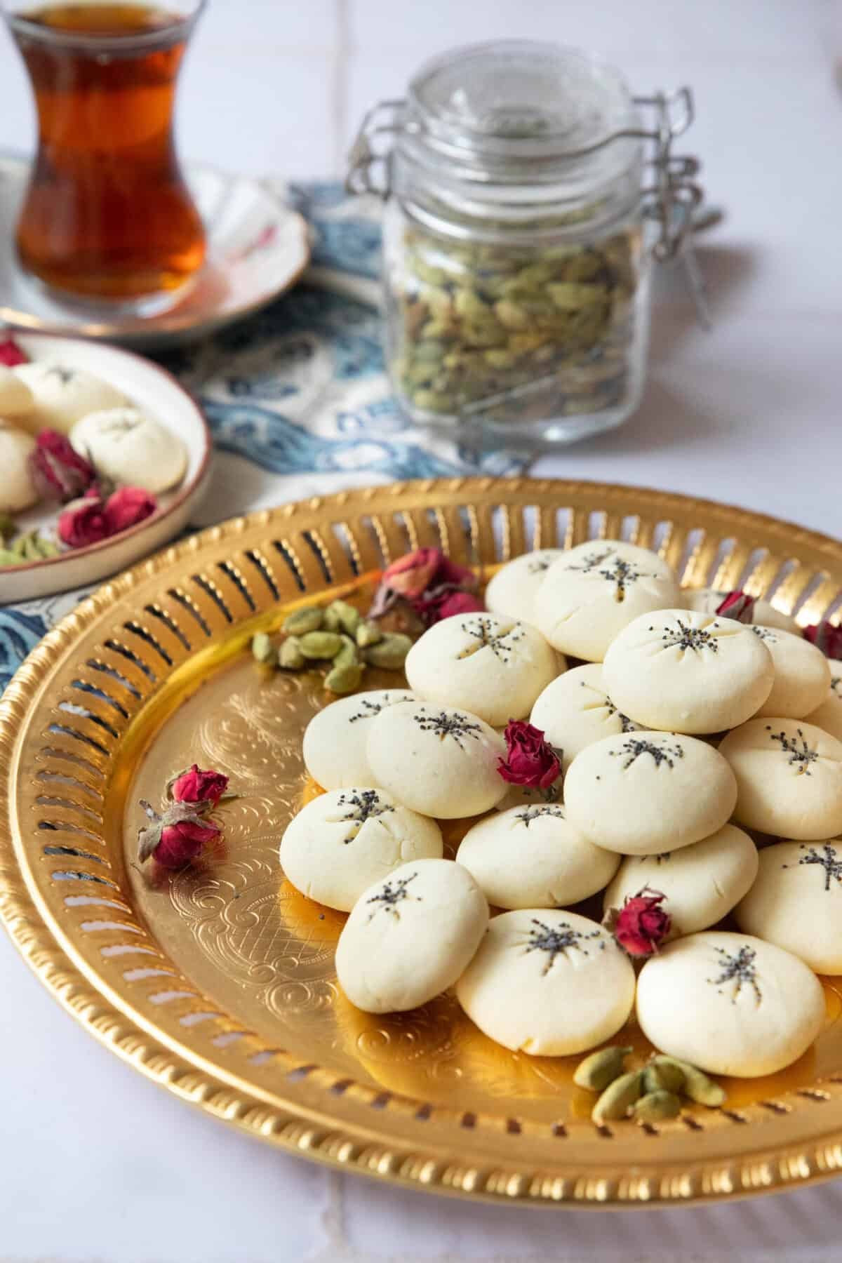 Persian Rice Cookies with Poppy Seeds a side view of nan e berenji cookies served on a copper tray with cardamom pods and rose petals alongside a teacup