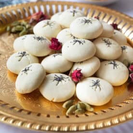 a close shot of nan e berenji cookies topped with poppy seeds and served on a copper tray next to cardamom pods and rose petals.