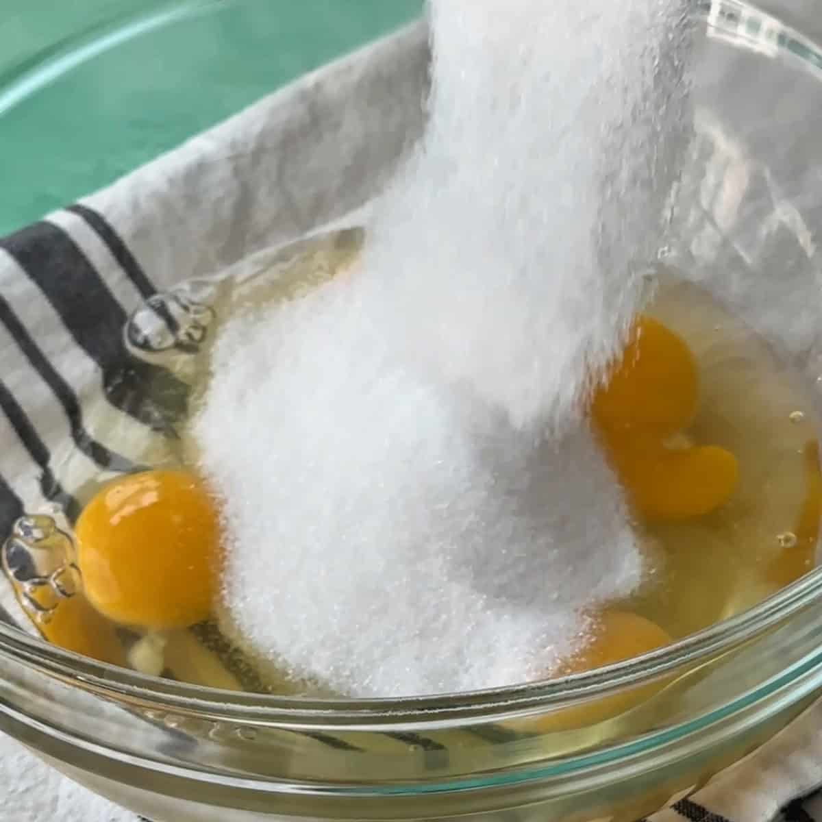 adding sugar to the cracked eggs in a transparent glass bowl