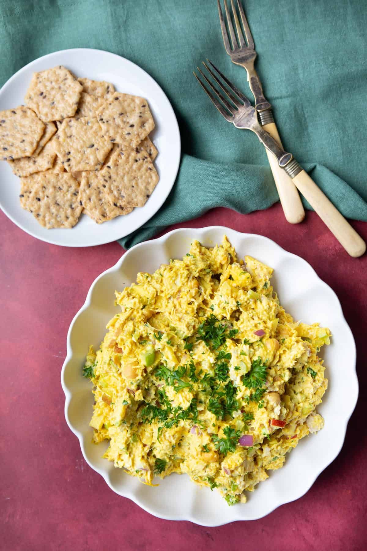 A bowl of chicken chickpea salad and a plate of crackers on a maroon backdrop